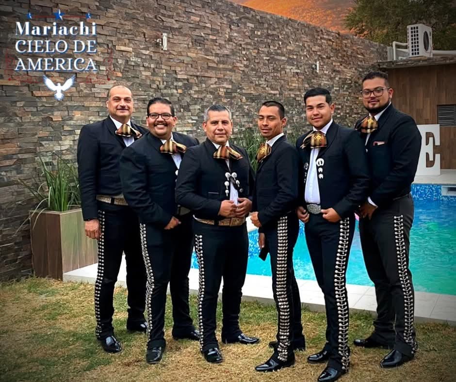 Los músicos de Mariachi Cielo de América, de traje negro con moño dorado, junto a una pared de piedra.